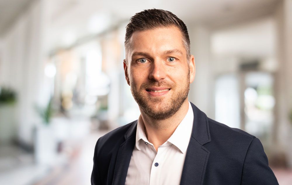 Freundliches Porträt von Dean Buhmeier, Ansprechpartner im Außendienst. Lächelnder Mann im Anzug, in einem modernen Büroambiente.